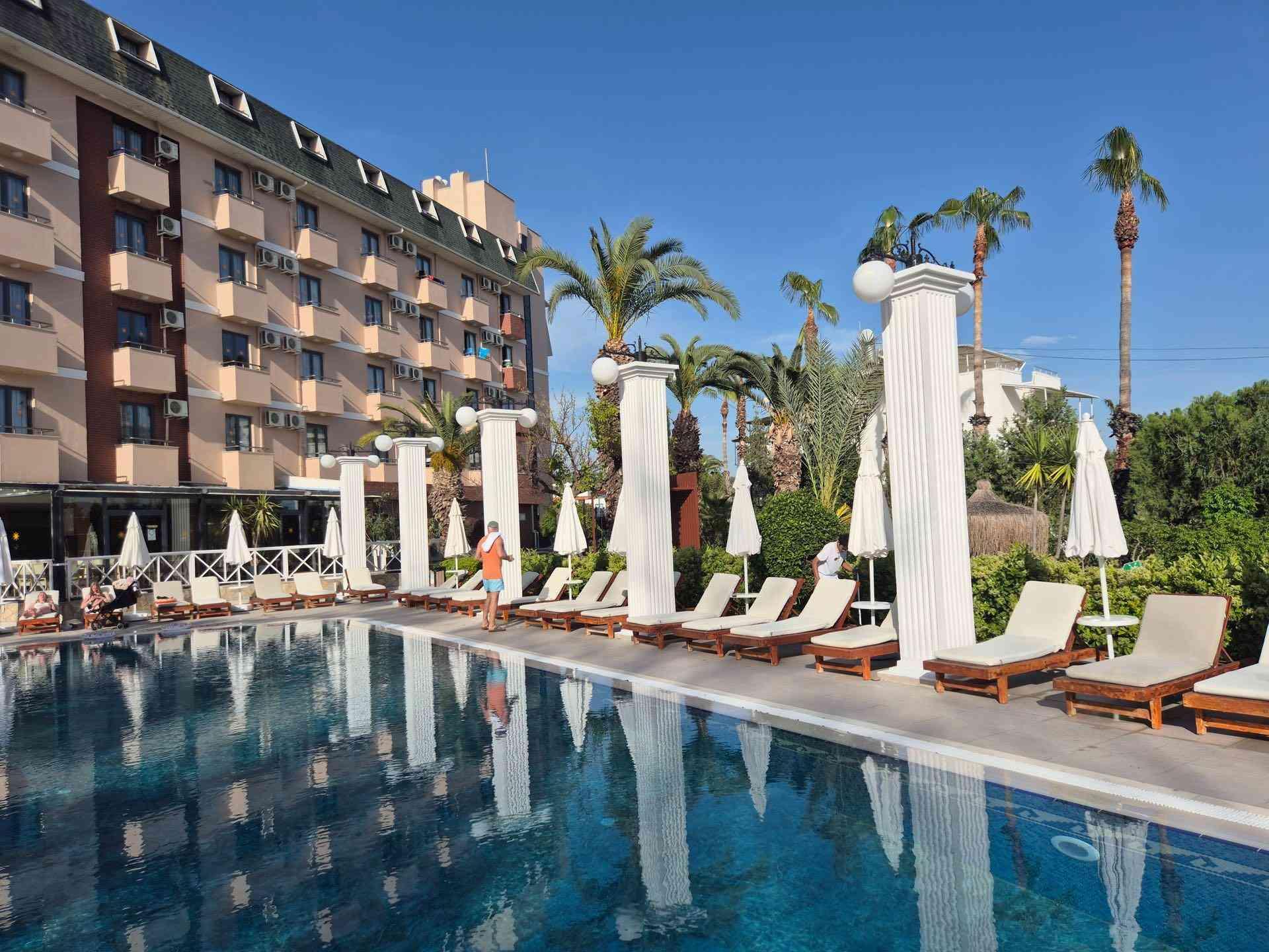 Moderner Hotelkomplex mit großem Pool, weißen Liegen, Sonnenschirmen und hohen Palmen bei klarem Himmel.