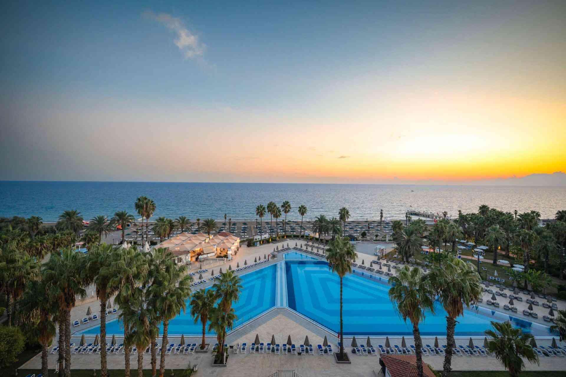 Großer Pool mit Palmen an der Küste, Sonnenuntergang am Meer, entspannte Hotel-Urlaubsatmosphäre.