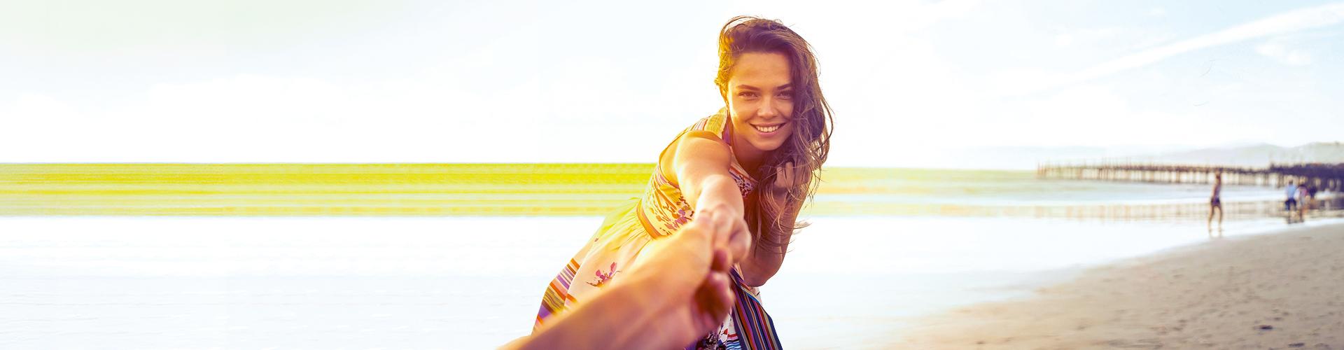 Glückliche Frau am sonnigen Strand, Hand haltend, mit weitem Blick aufs Meer und hellem Sand.