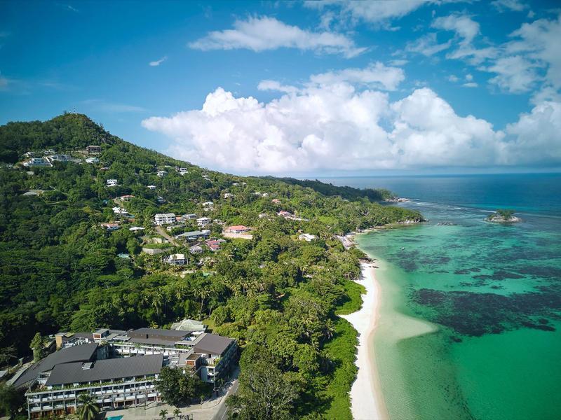 Hotelanlage am grünen Hang mit weißem Sandstrand und türkisfarbenem Meer unter blauem Himmel.