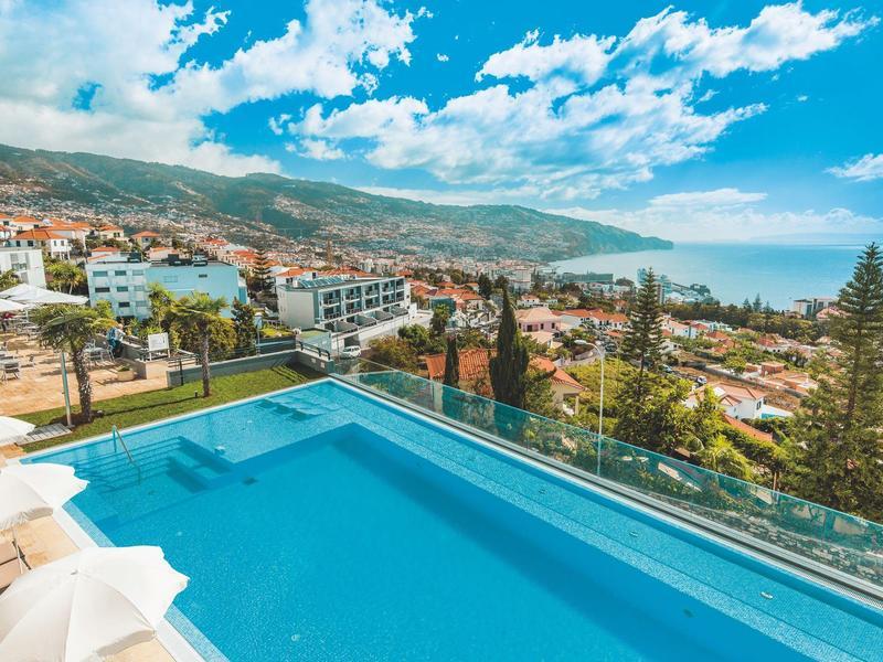 Moderner Hotelpool mit Glasgeländer, Blick auf Küste, Stadt und bewachsene Berge unter blauem Himmel.