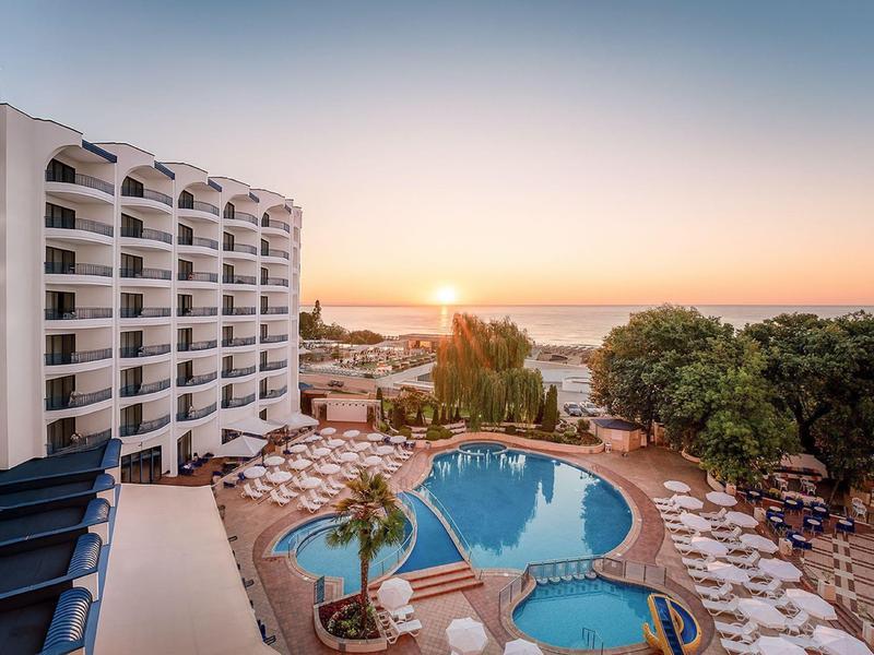 Großes Hotel mit Pool und Liegestühlen, Palmen und Sonnenuntergang am Meer im Hintergrund.