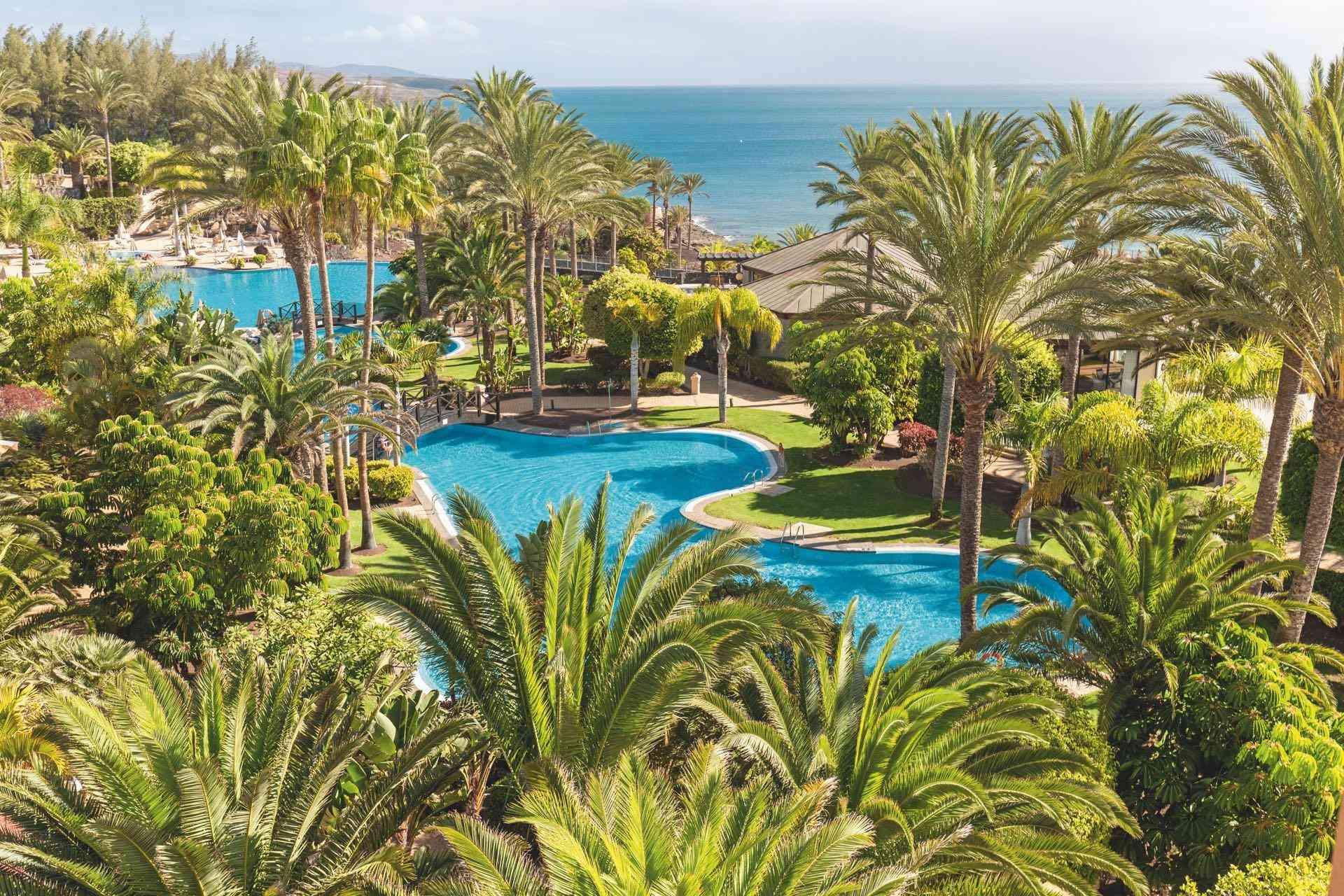 Luxuriöse Hotelanlage mit mehreren Pools, Palmen und Blick auf das blaue Meer am Horizont.