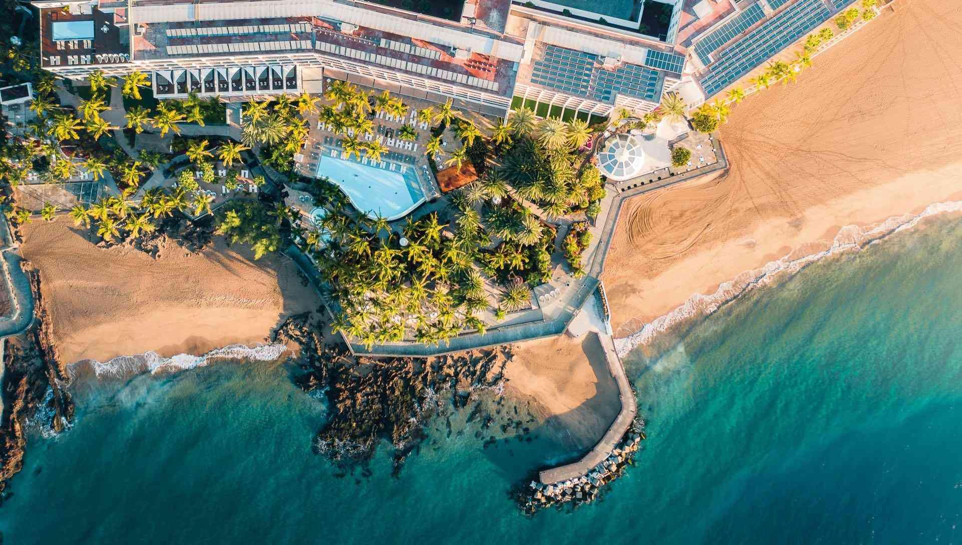 Luxushotel mit Pool, Palmen und Strand an türkisfarbenem Meer, ideal für entspannte Ferien.