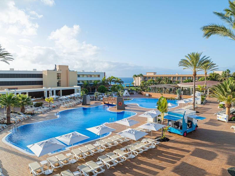 Moderner Hotelpoolbereich mit Sonnenschirmen, zahlreichen Liegestühlen und Palmen unter blauem Himmel.