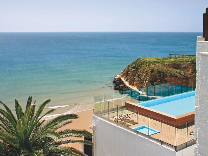 Infinity-Pool mit Glasgeländer über Meer und Sandstrand, Palmen und felsige Küste unter klarem Himmel.