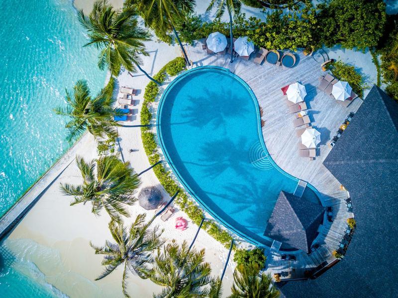 Ein tropischer Hotelpool mit Palmen, Sonnenschirmen und türkisblauem Meer im Hintergrund.