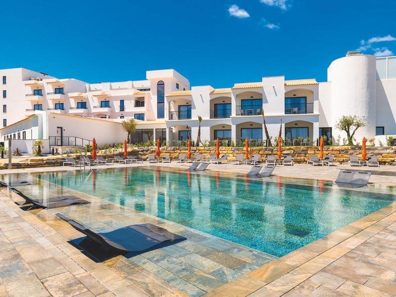 Modernes Hotel mit weißer Fassade, großem Pool und Liegestühlen unter strahlend blauem Himmel.