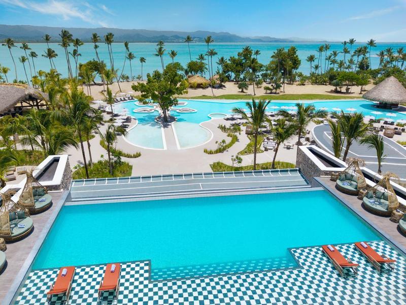 Ein großer Hotelpool mit Liegestühlen, Palmen und Meerblick unter blauem Himmel lädt zum Entspannen ein.
