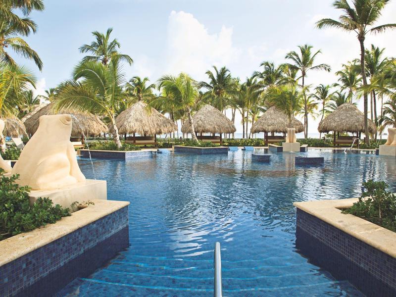 Großer Pool mit klar blauem Wasser, umgeben von Palmen und Strohdächern in tropischer Hotelanlage.