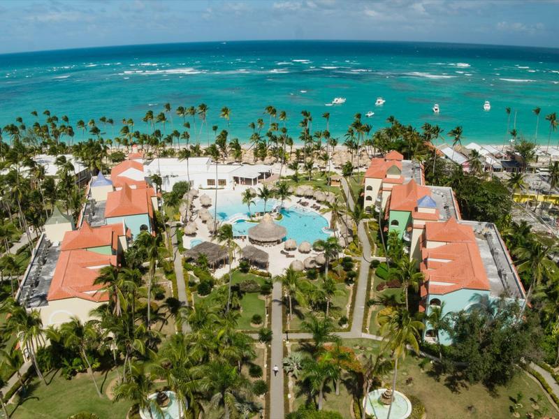 Luxuriöses Hotel mit roten Dächern, Pool, Palmen und weißem Sandstrand am türkisfarbenen Meer.