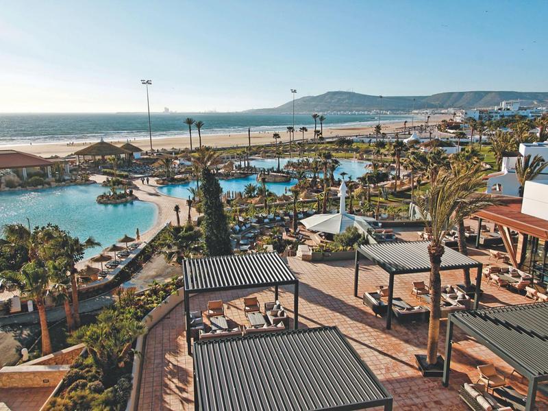 Hotelanlage mit mehreren Pools, Liegestühlen, Palmen und weitem Blick auf den Sandstrand und das Meer.