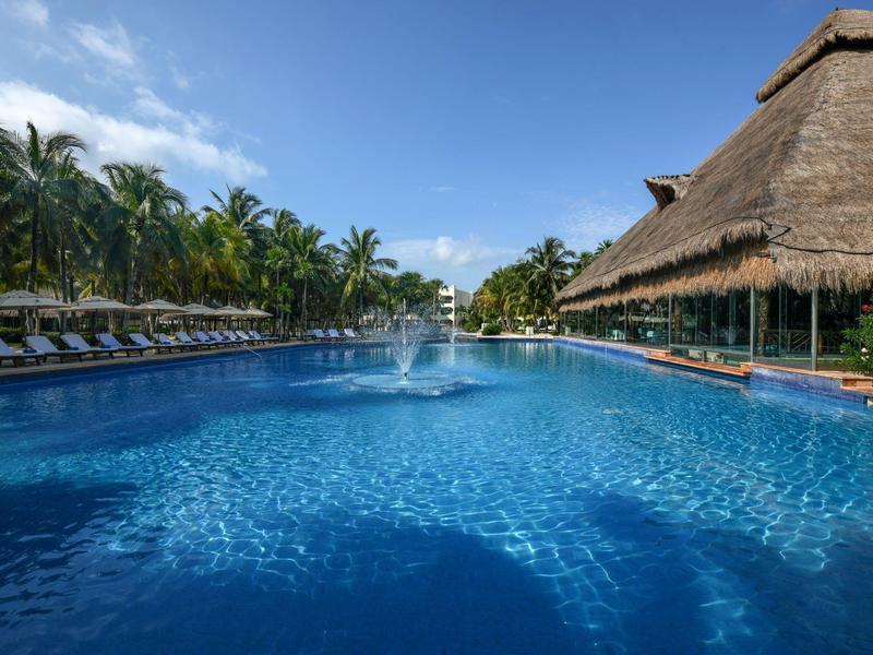 Großer Hotelpool mit Fontäne, umgeben von Palmen, Liegen und Strohdach unter blauem Himmel.