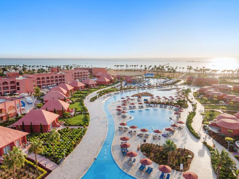 Großes Hotelresort mit Poollandschaft, roten Dächern, grünen Gärten und Meer am Horizont bei Sonnenaufgang.