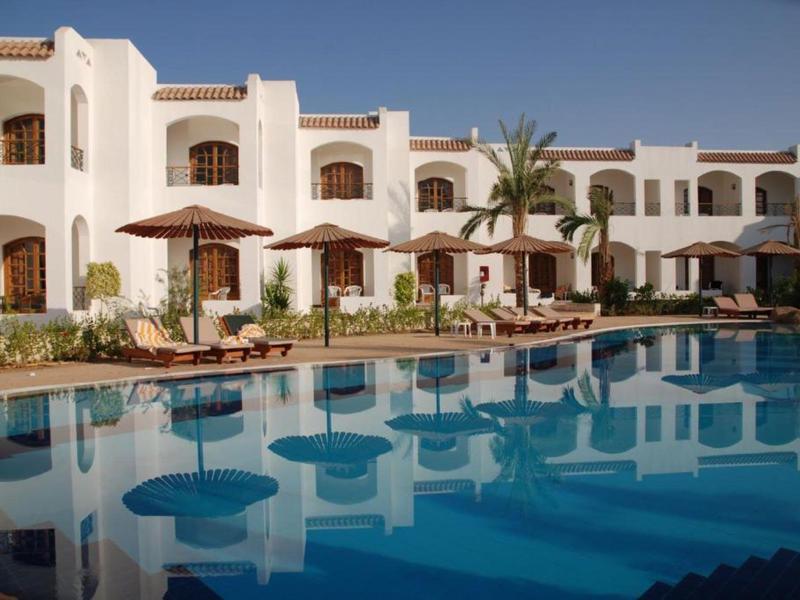 Ein Hotel mit weißen Gebäuden und braunen Sonnenschirmen neben einem großen, klaren Pool bei blauem Himmel.