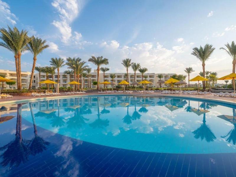 Großer Pool mit gelben Schirmen, Palmen und modernem Hotelgebäude bei klarem Himmel.
