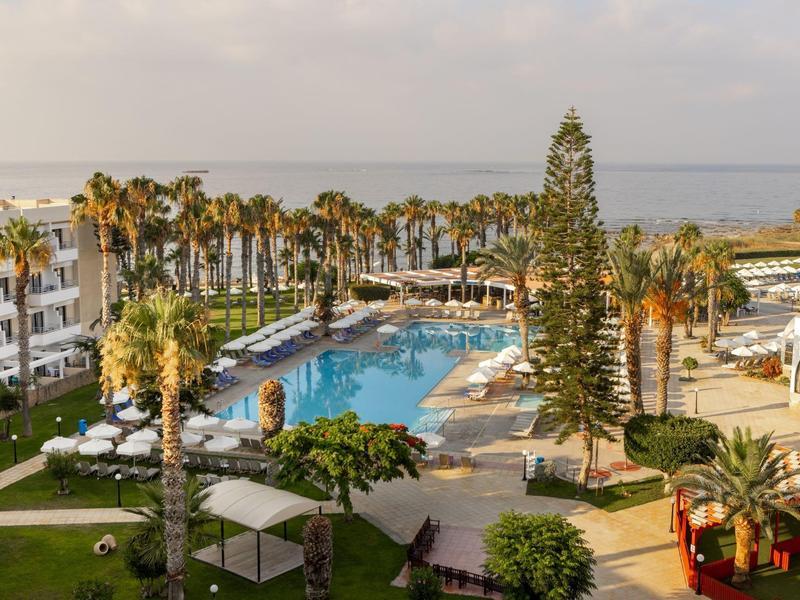 Hotelanlage mit großem Pool, Palmen, Liegen und Meer im Hintergrund bei klarem Himmel.