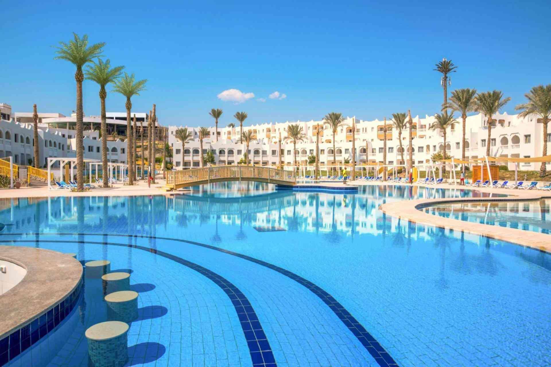 Großer Hotelpool mit klarblauem Wasser, Palmen und weißem Gebäude unter klarem Himmel.