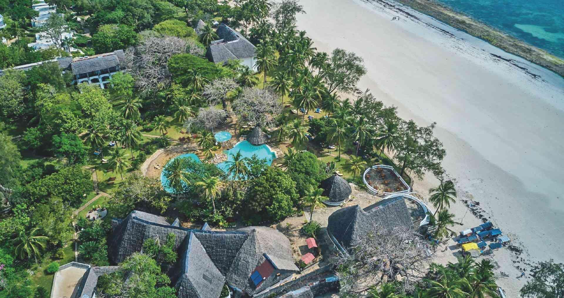 Hotelanlage mit Pool, grüner Vegetation und langem Sandstrand neben klarem Meer.