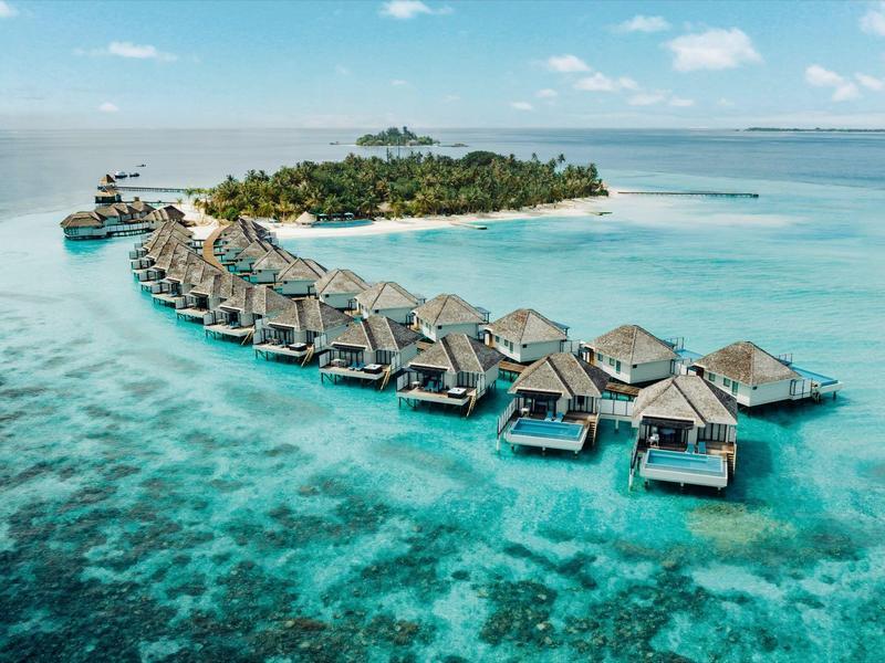 Luxuriöse Überwasserbungalows in türkisblauem Wasser vor einer tropischen Insel mit Palmen.