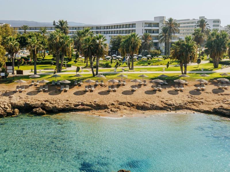 Hotelanlage mit grüner Palmenlandschaft, Sonnenliegen am Sandstrand und klarem türkisfarbenem Meer.