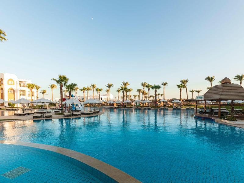 Großer blauer Hotelpool mit Palmen, Sonnenschirmen und Relaxbereichen unter klarem Himmel.