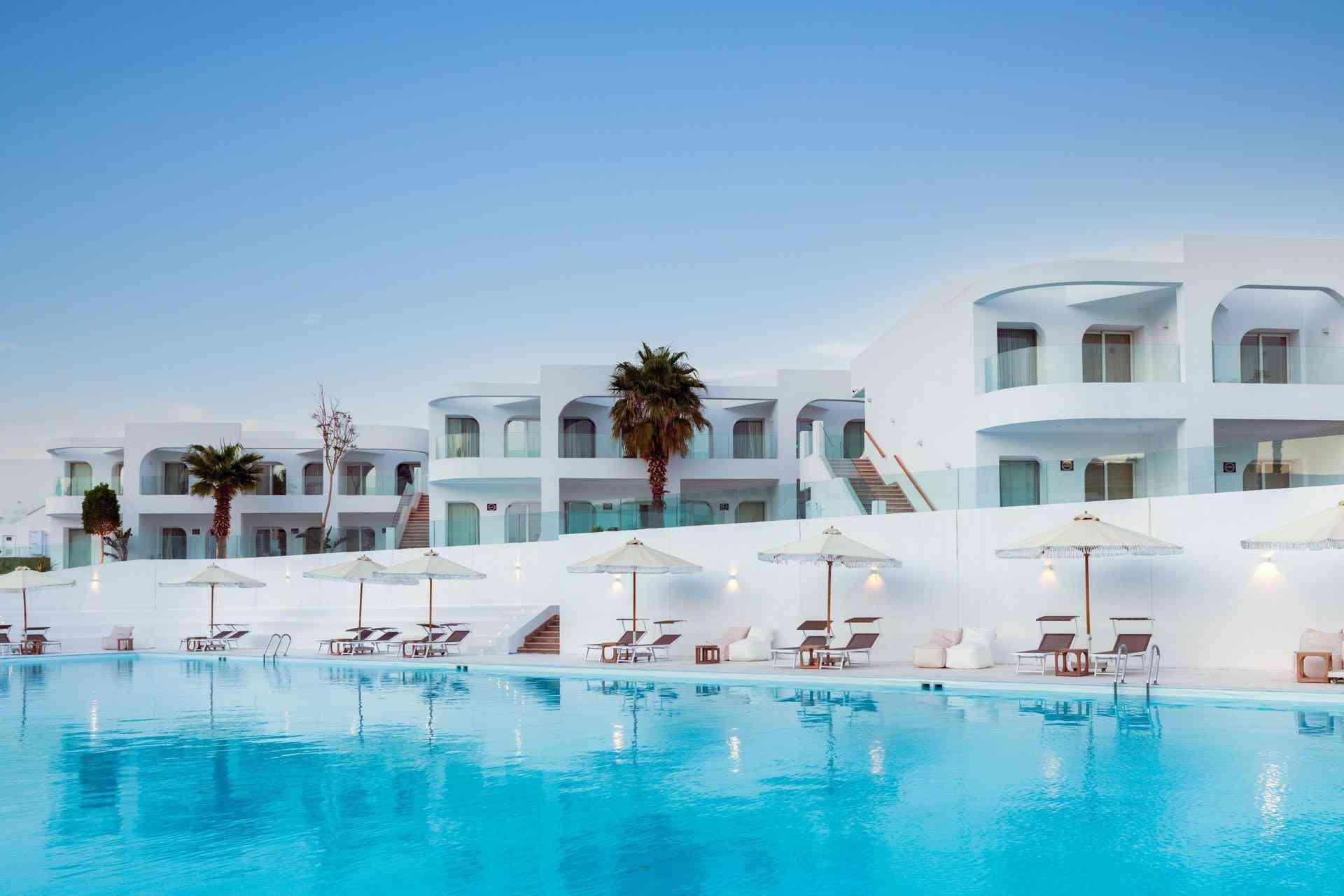 Luxuriöses Hotel mit weißer Fassade und großem Pool, umgeben von Sonnenschirmen und Palmen.