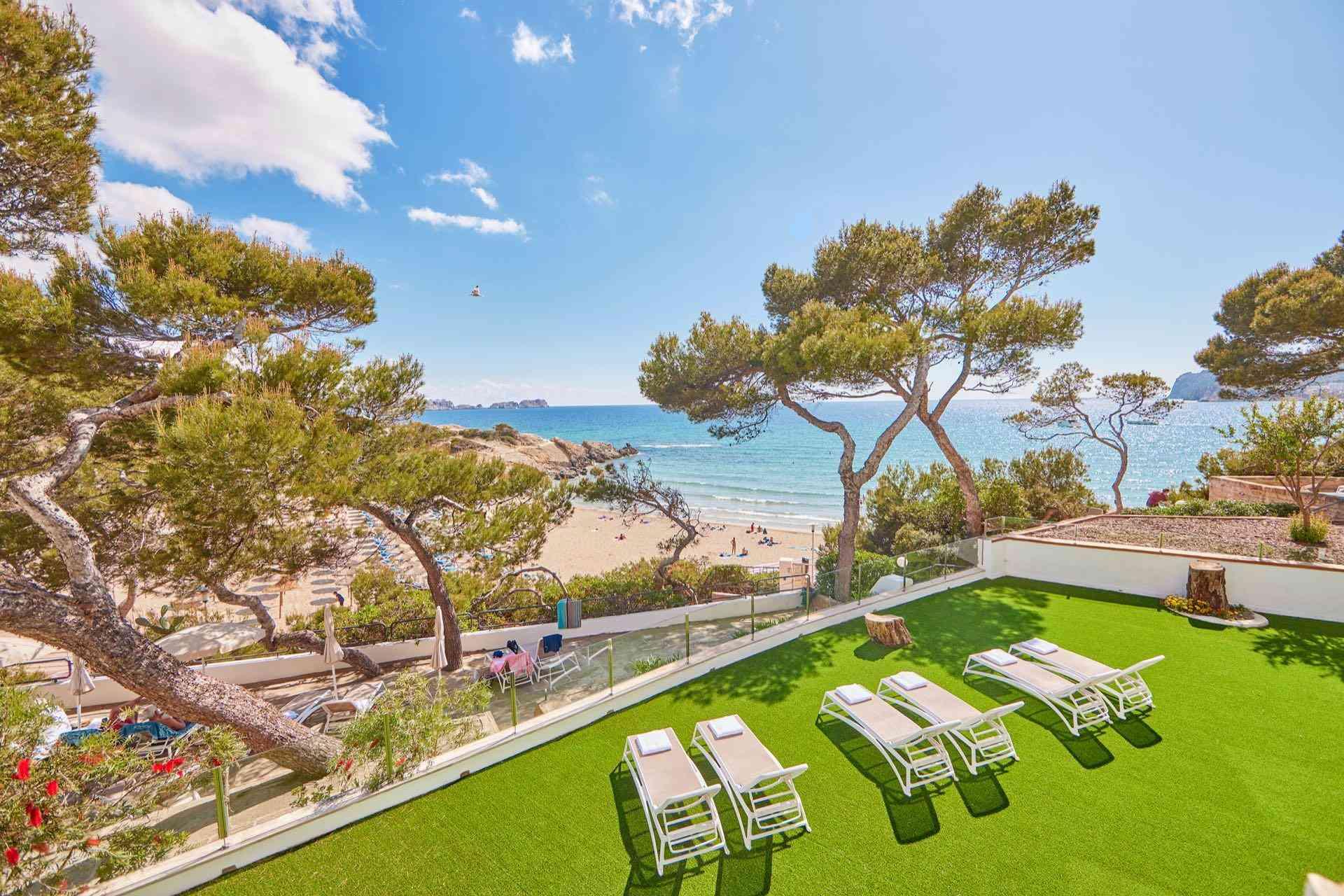 Hotelterrasse mit Liegestühlen, Pinienbäumen und Blick auf den Sandstrand und das blaue Meer.