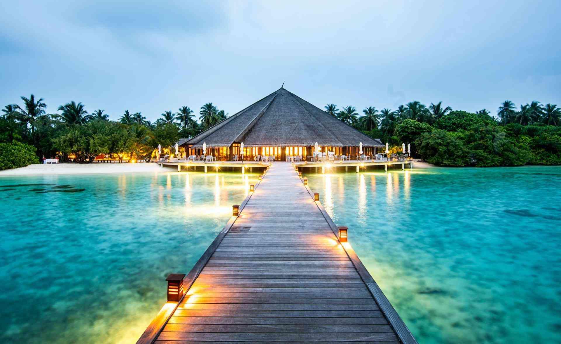 Luxusresort mit beleuchtetem Steg und Palmen am türkisfarbenen, klaren Wasser bei Abenddämmerung.