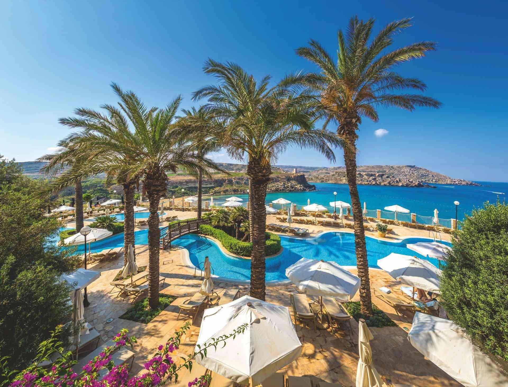 Luxuriöse Hotelpoolanlage mit Palmen, Sonnenschirmen und Blick auf das blaue Meer unter klarem Himmel.
