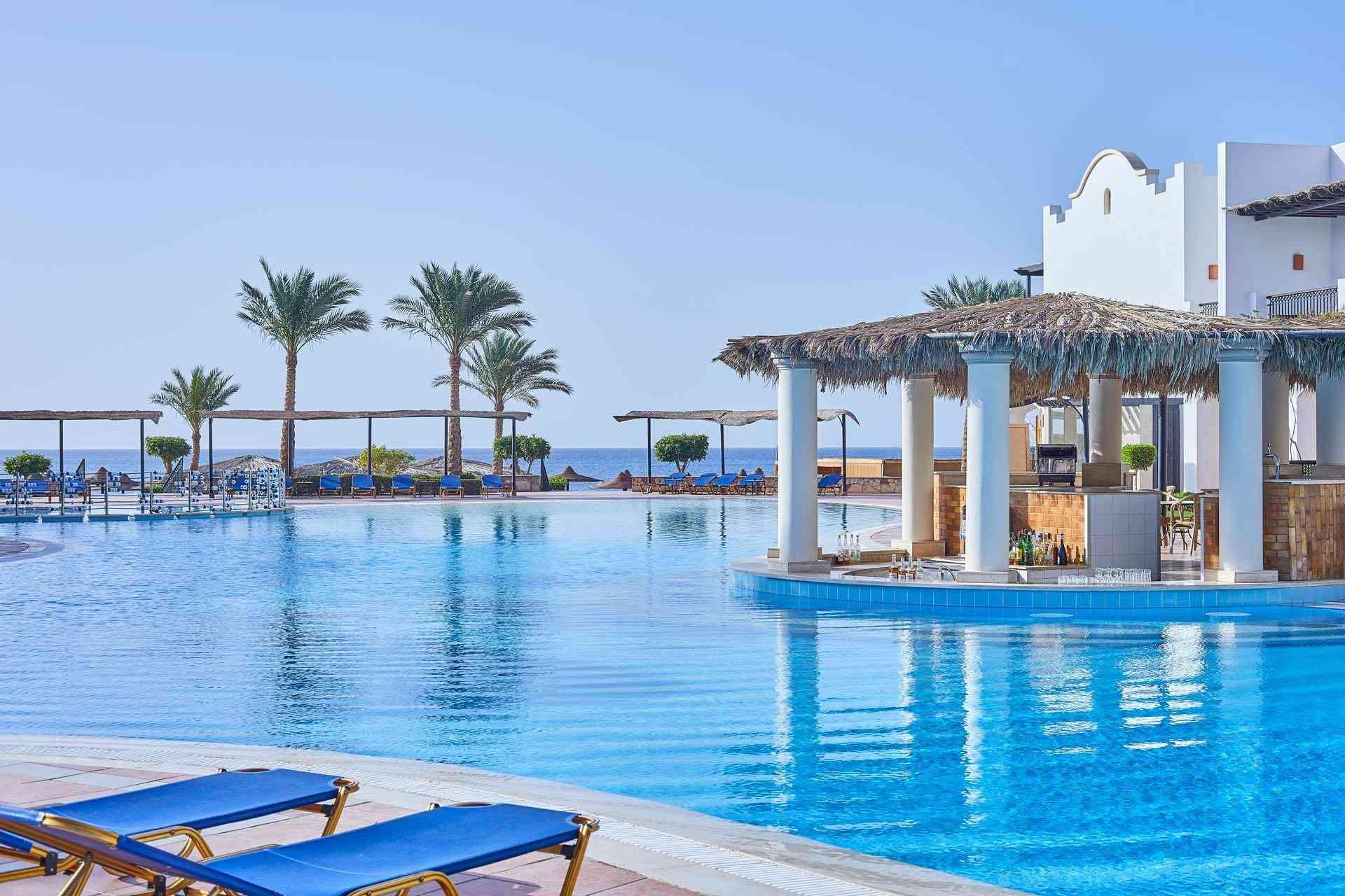 Großer Hotelpool mit integriertem Barbereich, blauer Liegestühle und Palmen am Meer im Hintergrund.