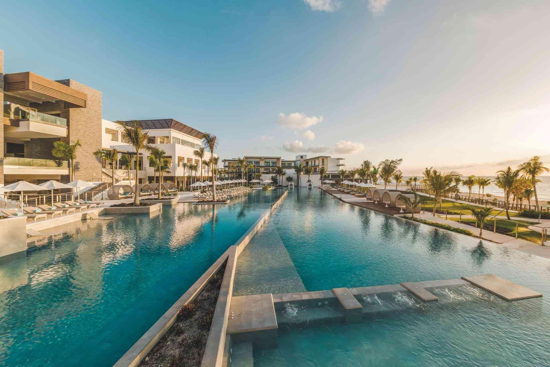 Luxuriöses Hotel mit großem, klaren Pool, Palmen und komfortablen Liegen unter blauem Himmel.