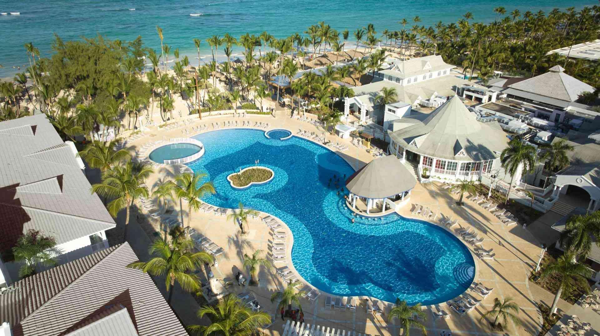 Luxuriöses Hotel mit großem geschwungenem Pool, Palmen, Sonnenschirmen und Strand am türkisblauen Meer.