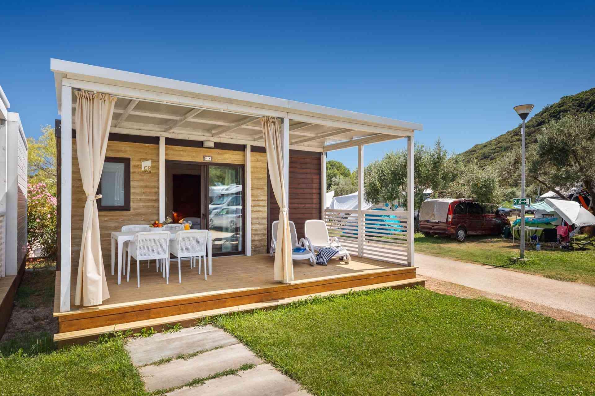 Kleines modernes Ferienhaus mit überdachter Terrasse, Garten und Liegestühlen bei sonnigem Wetter.