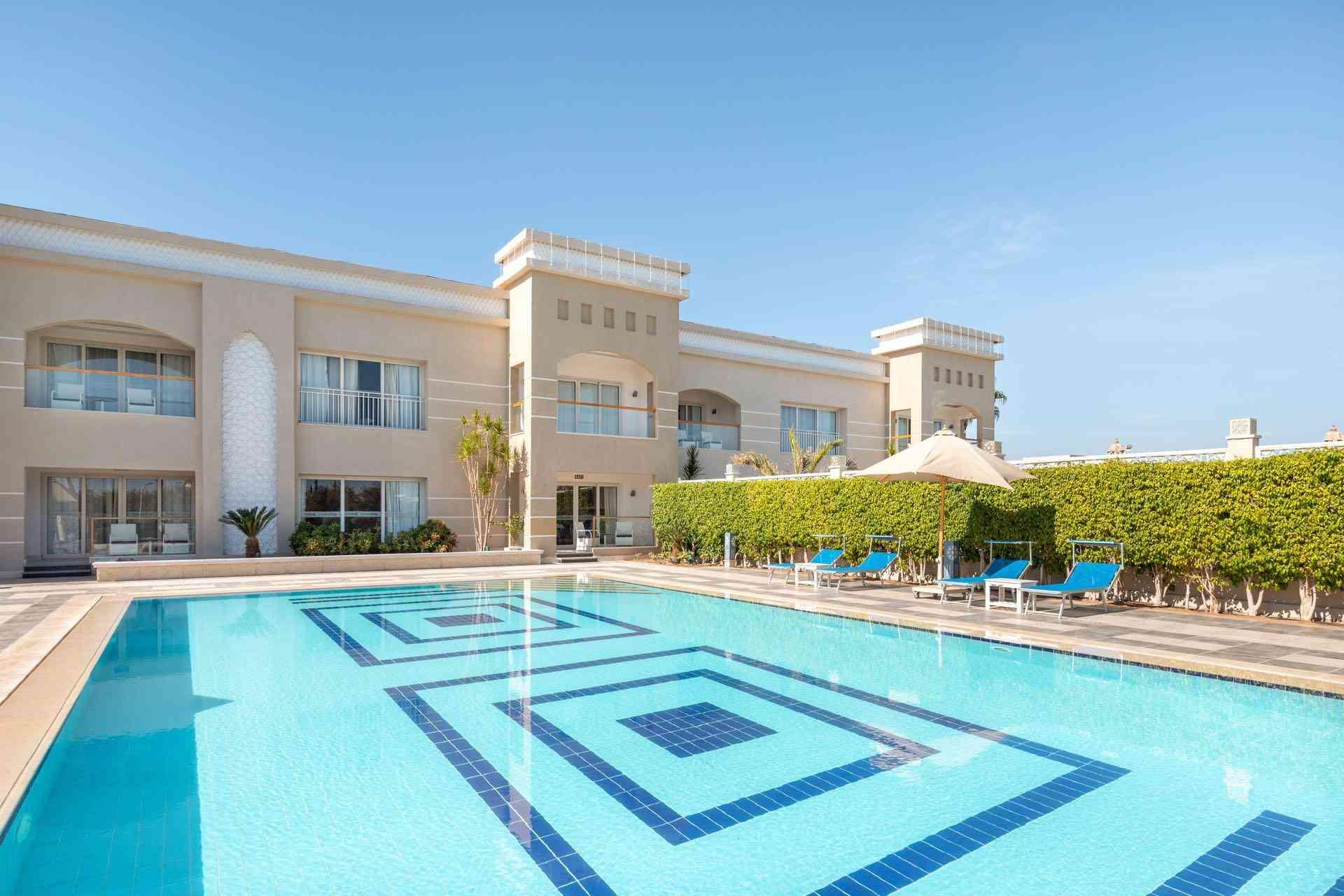 Moderner Hotelpool mit blauem Wasser, Liegestühlen und beige-weißer Fassade bei klarem Himmel.