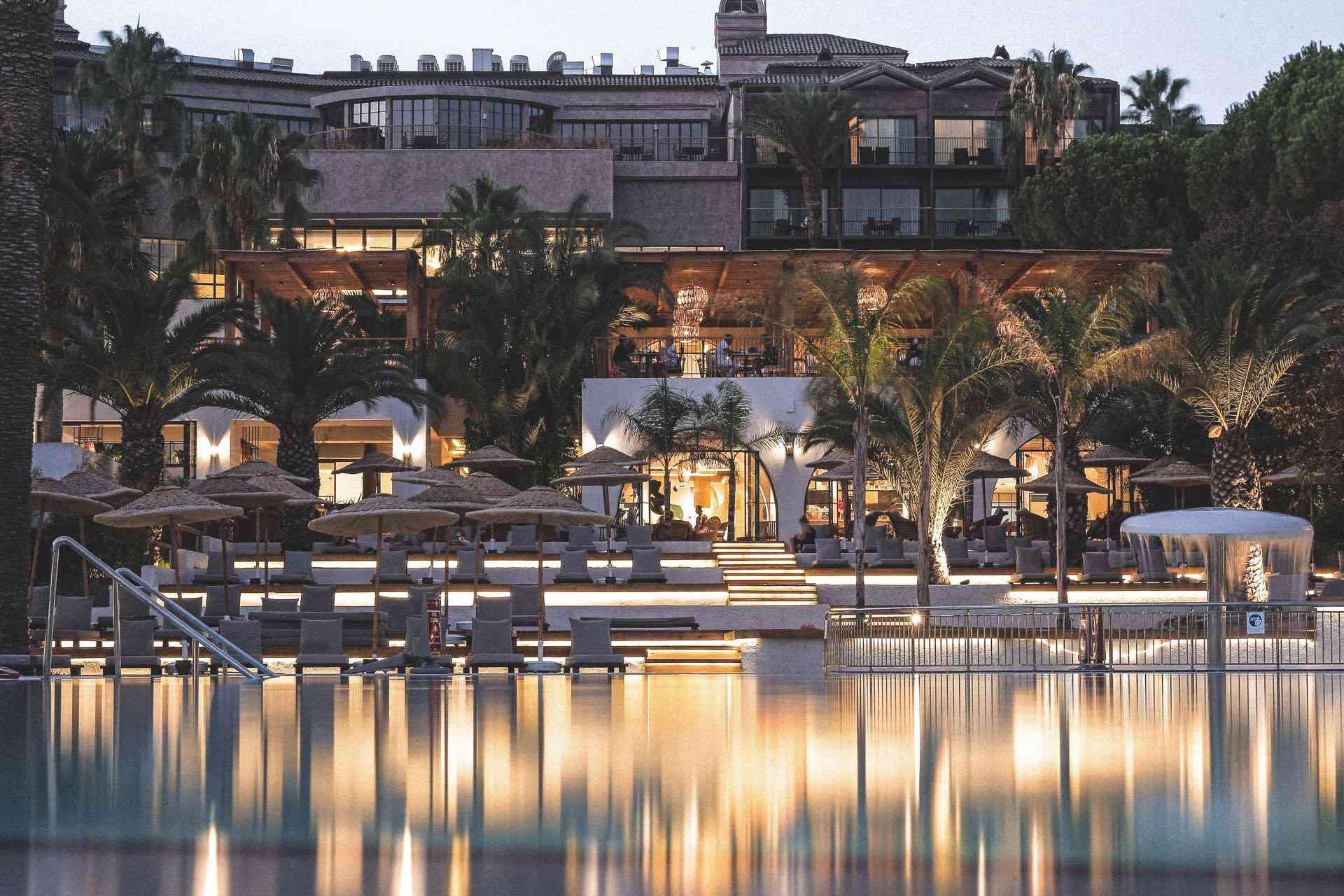 Luxuriöses Hotel mit beleuchtetem Pool, Liegen, Palmen und mediterraner Architektur bei Abenddämmerung.