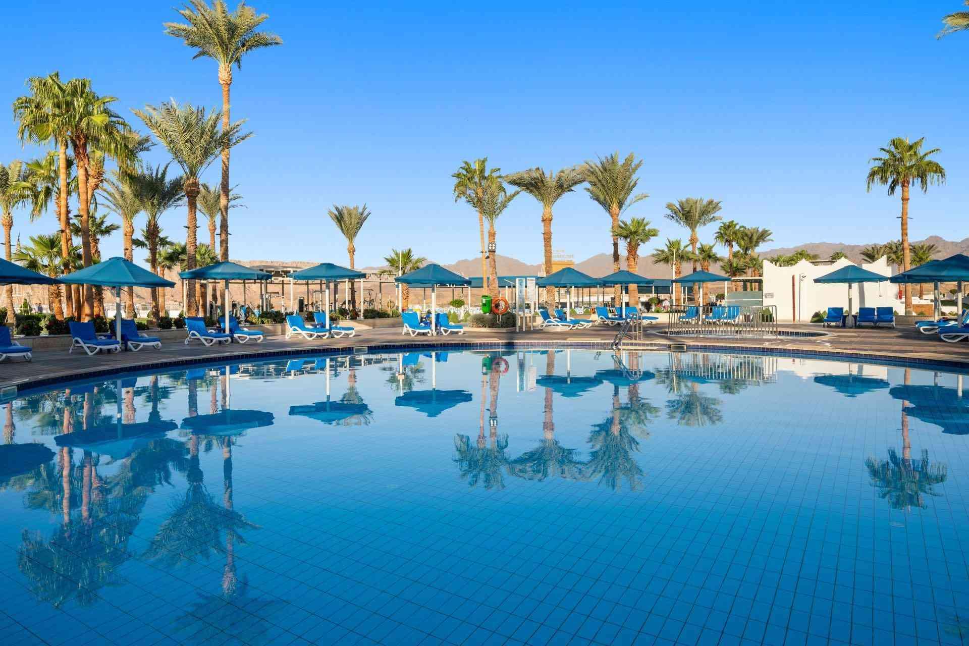 Großer Hotelpool mit blauer Wasseroberfläche, umgeben von Sonnenschirmen und Palmen unter blauem Himmel.