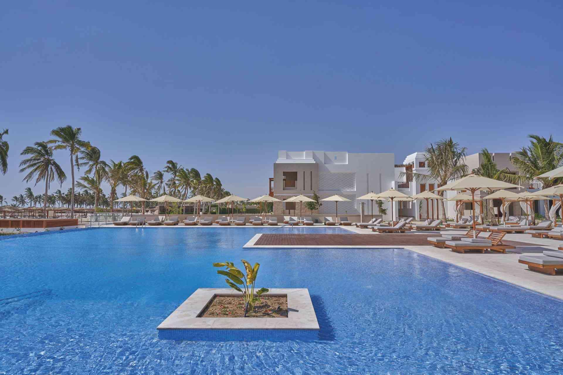 Großer blauer Hotelpool mit Liegen, Sonnenschirmen und Palmen unter klarem Himmel.