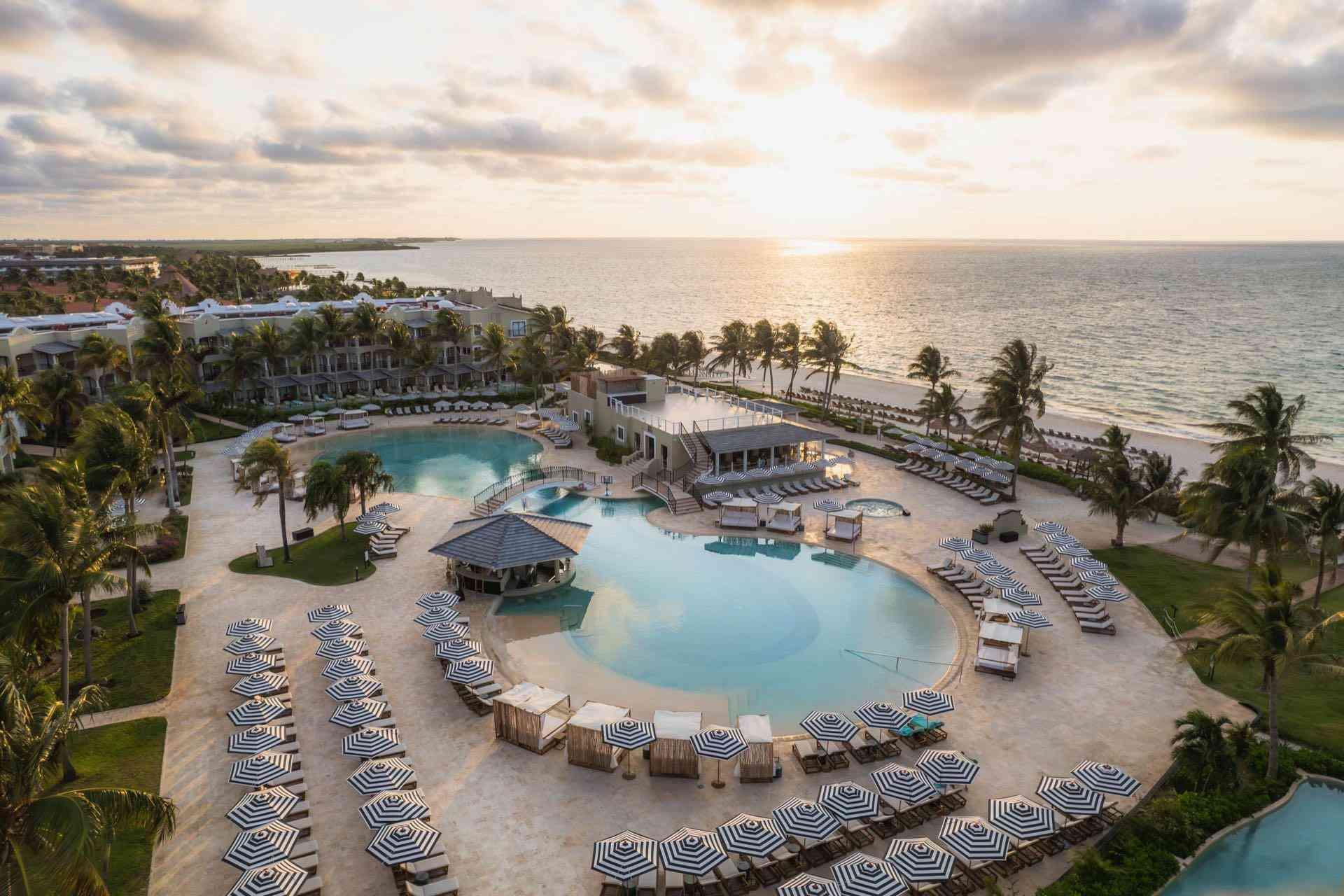Hotelresort mit großem Pool, Sonnenliegen, Palmen und Blick auf ruhiges Meer bei Sonnenuntergang.