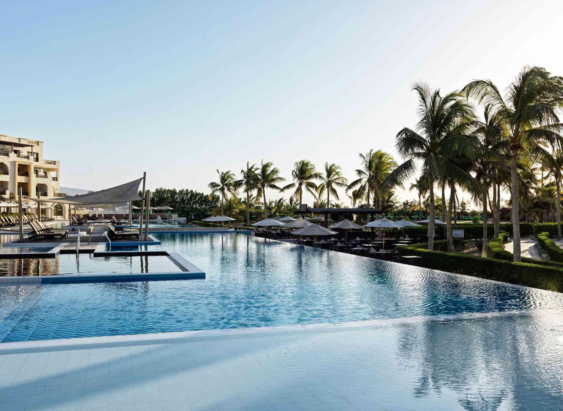 Großer Hotelpool mit Palmen, Liegestühlen und Blick auf Gebäude bei klarem Himmel.