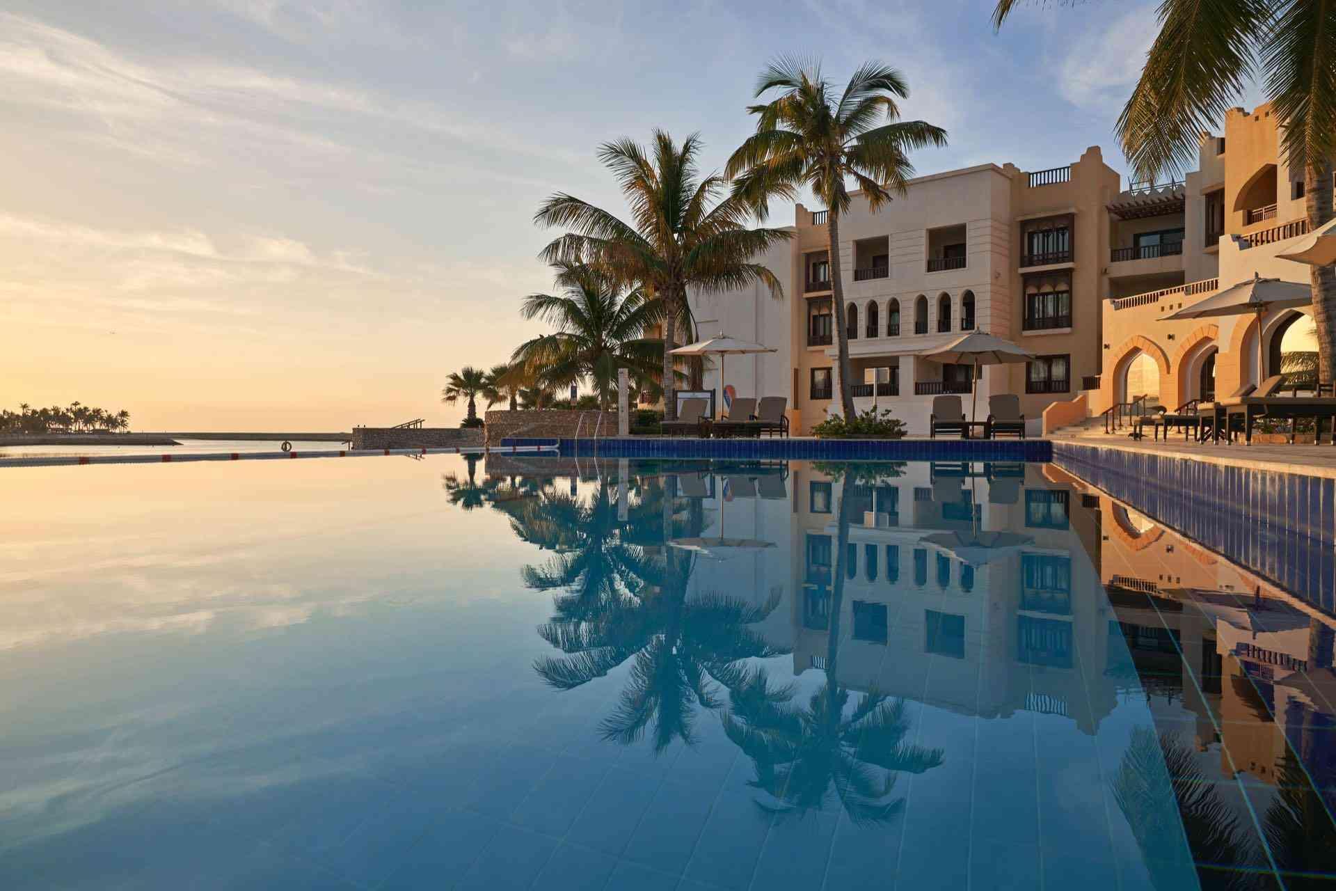Hotel mit großem Pool und Palmen bei Sonnenuntergang, ruhige Urlaubsatmosphäre am Wasser.