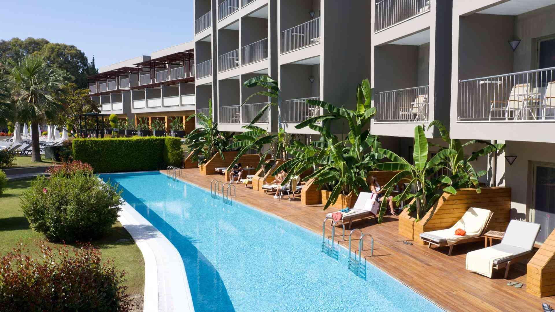 Moderner Hotelpool mit Liegestühlen und Balkonen, umgeben von tropischer Bepflanzung.