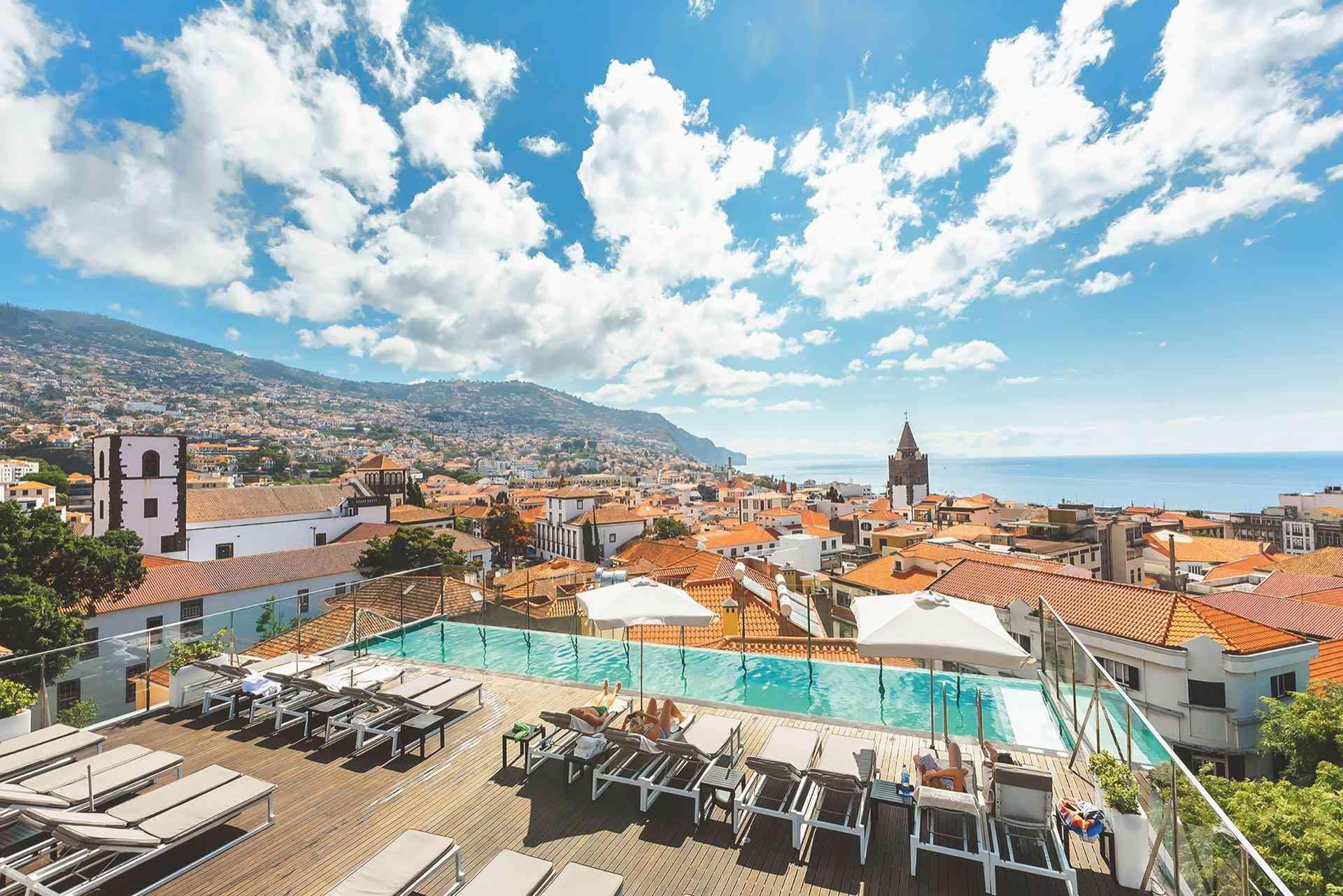 Hotel mit Infinity-Pool, Liegestühlen und Meerblick in sonniger Küstenstadt mit Bergen und Wolken.