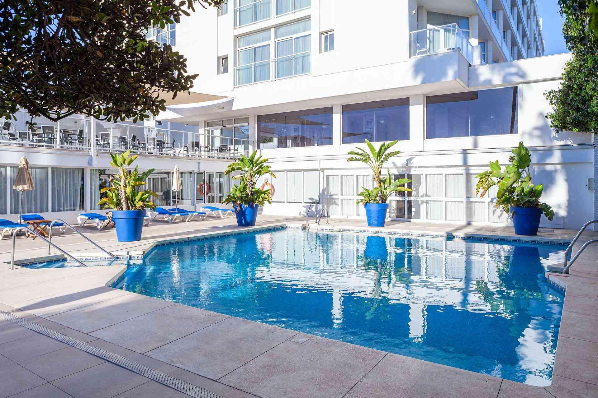 Moderner Hotelpool mit blauen Pflanzenkübeln, Sonnenliegen und weißem Gebäude im Hintergrund.