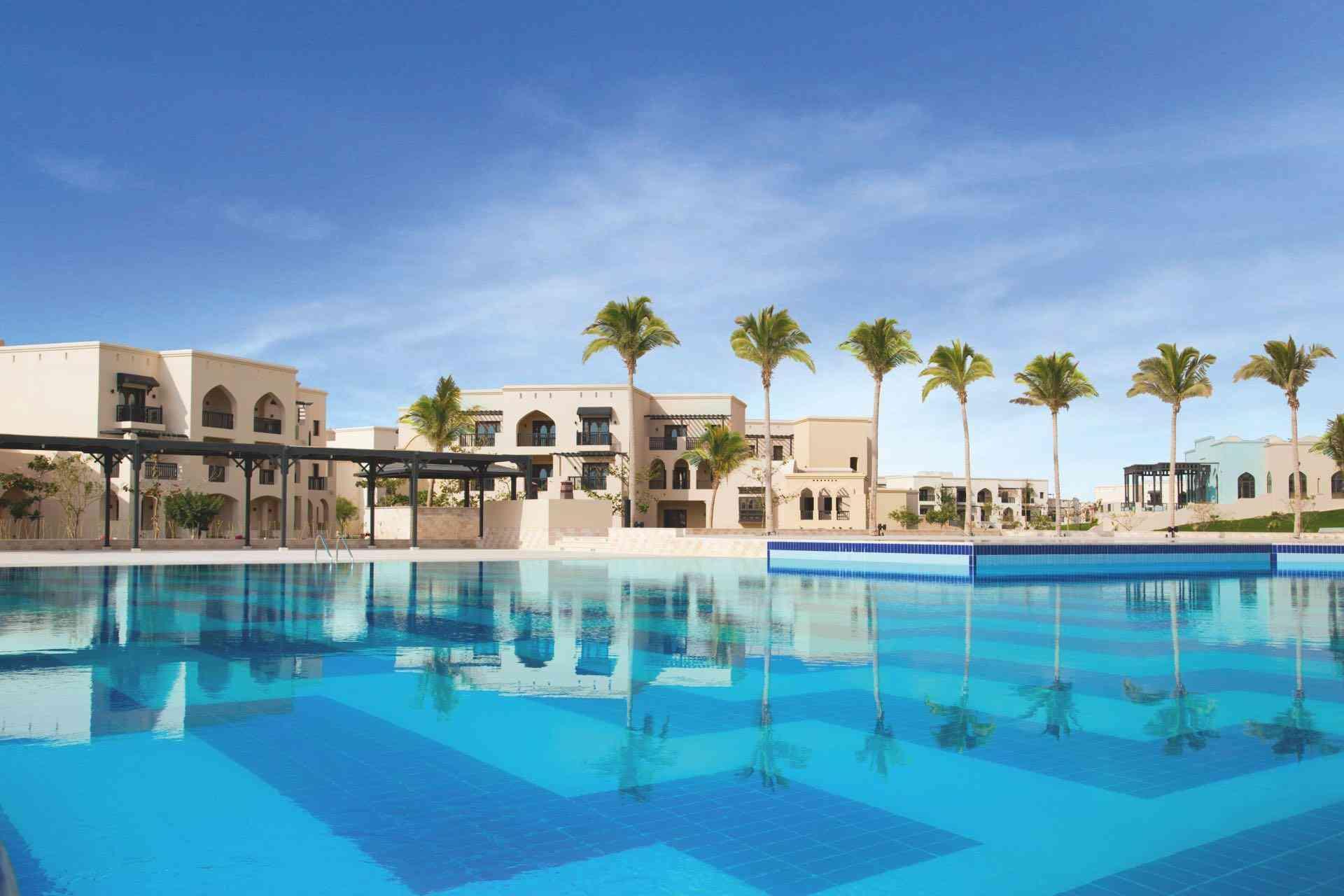 Moderner Hotelkomplex mit großem blauen Pool, Palmen und klarem Himmel für Urlaubsentspannung.