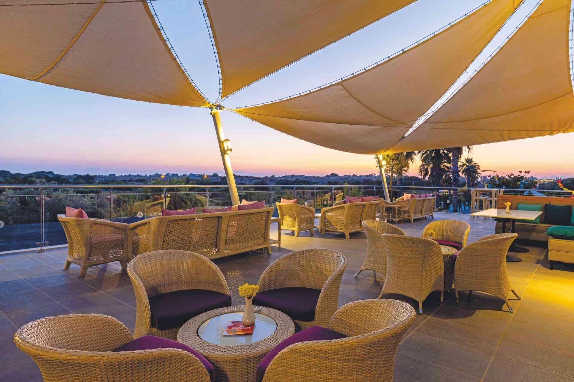 Offene Hotelterrasse mit bequemen Korbsesseln und Sonnensegeln bei Sonnenuntergang.