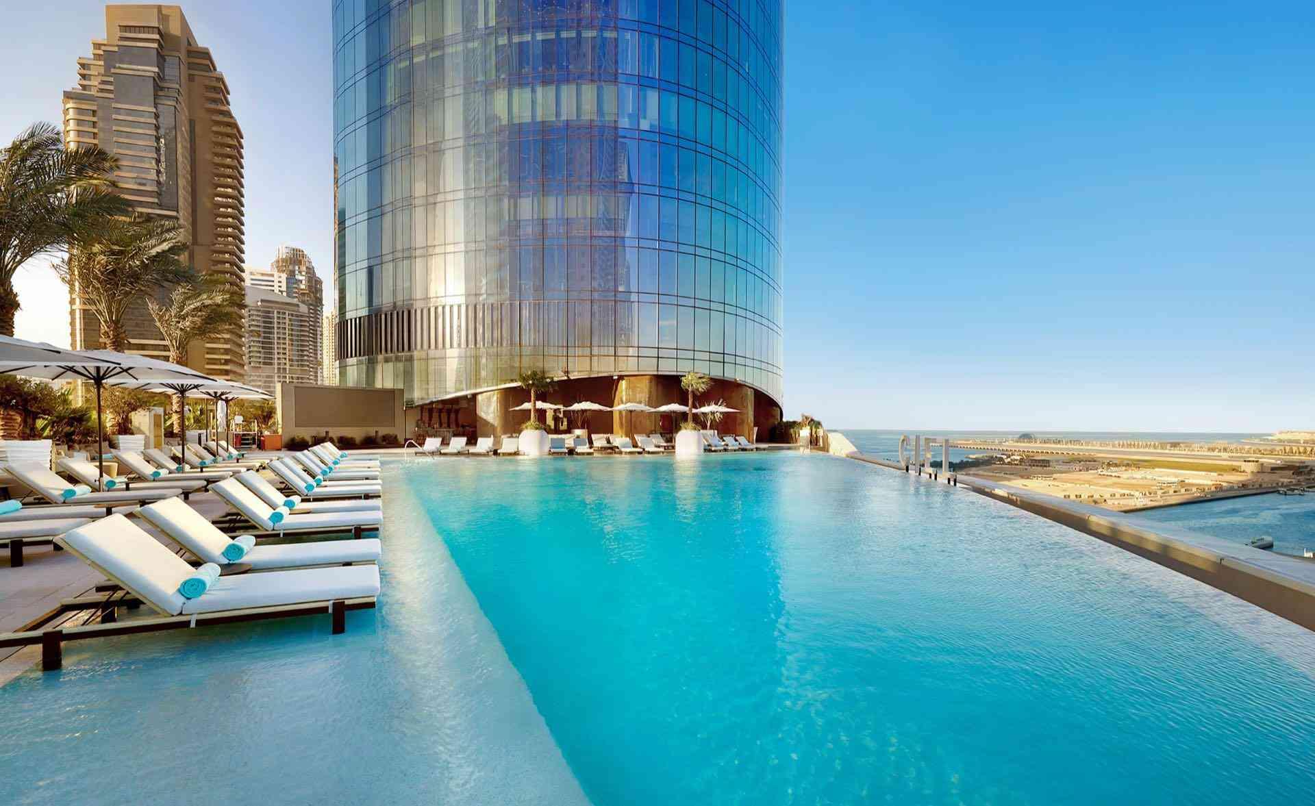 Infinity-Pool vor einem modernen Glas-Hochhaus mit Sonnenliegen und Blick auf eine Stadt- und Wüstenlandschaft.