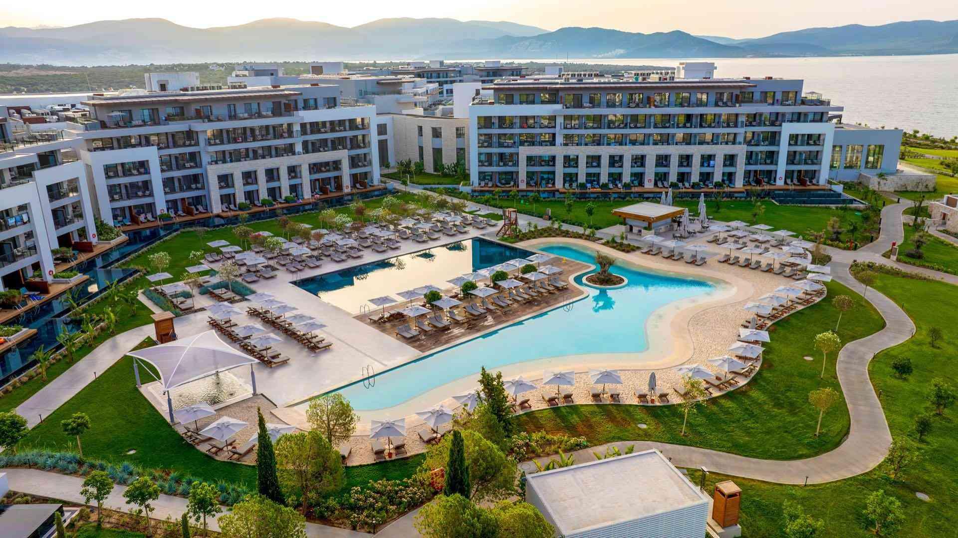 Luxuriöses Hotel mit großem Pool, Liegestühlen und Blick auf ruhiges Meer und Berge.