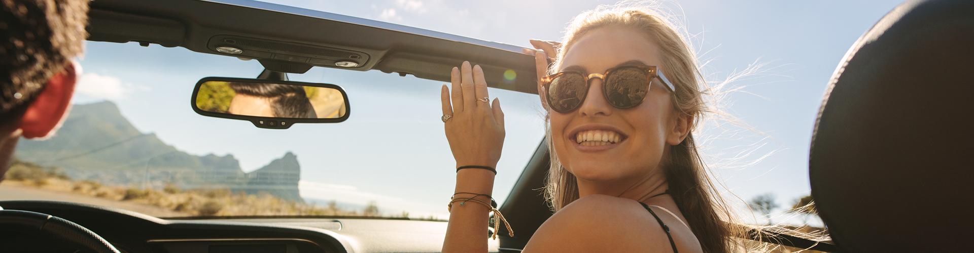 Glückliche Frau mit Sonnenbrille im Cabrio unterwegs, Sonne und Bergkulisse im Hintergrund.