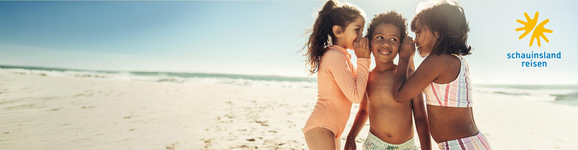 Drei lachende Kinder spielen am sonnigen Sandstrand, entspannte Urlaubsstimmung am Meer.